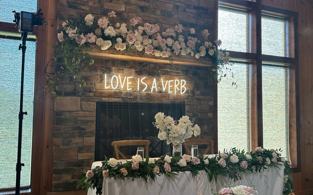 "Love is a Verb" in white neon lights, hanging above a fireplace behind a wedding sweetheart table adorned with white roses & greenery.